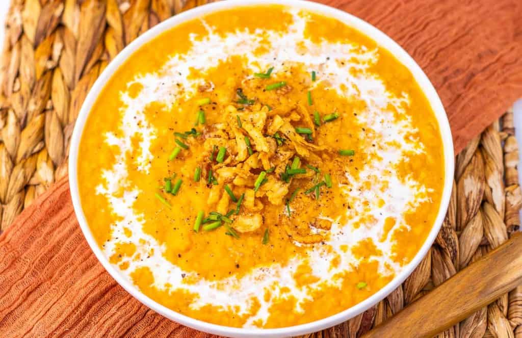 A bowl of creamy orange soup garnished with crispy onions, chopped herbs, black pepper, and a swirl of cream, placed on a woven mat with an orange napkin beside it.