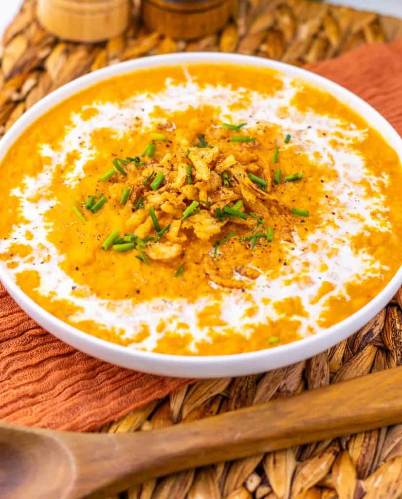 A bowl of creamy orange soup garnished with crispy fried onions, chopped chives, and a swirl of cream, placed on a woven mat next to a wooden spoon.