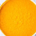 Overhead view of a pot filled with bright orange soup or stew, possibly containing blended vegetables or lentils.