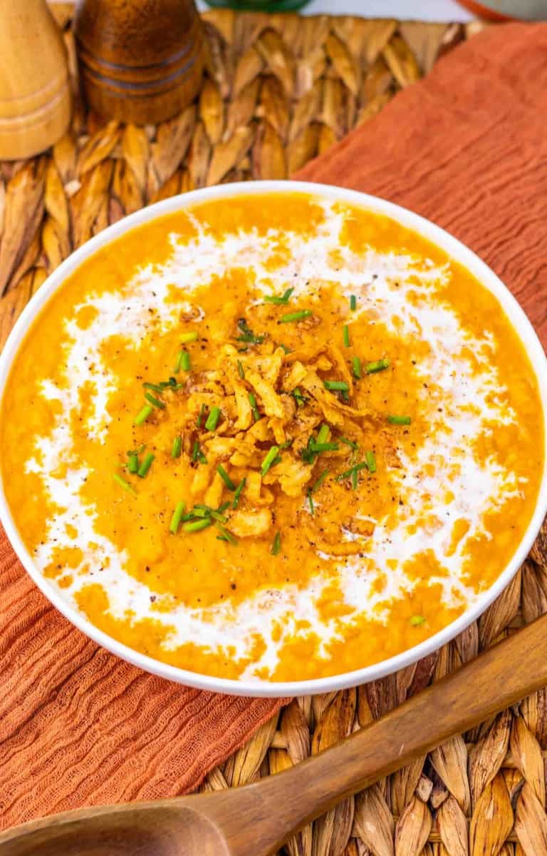 A bowl of yellow lentil dal topped with fried onions, herbs, and a swirl of cream, placed on a woven mat with a wooden spoon nearby.