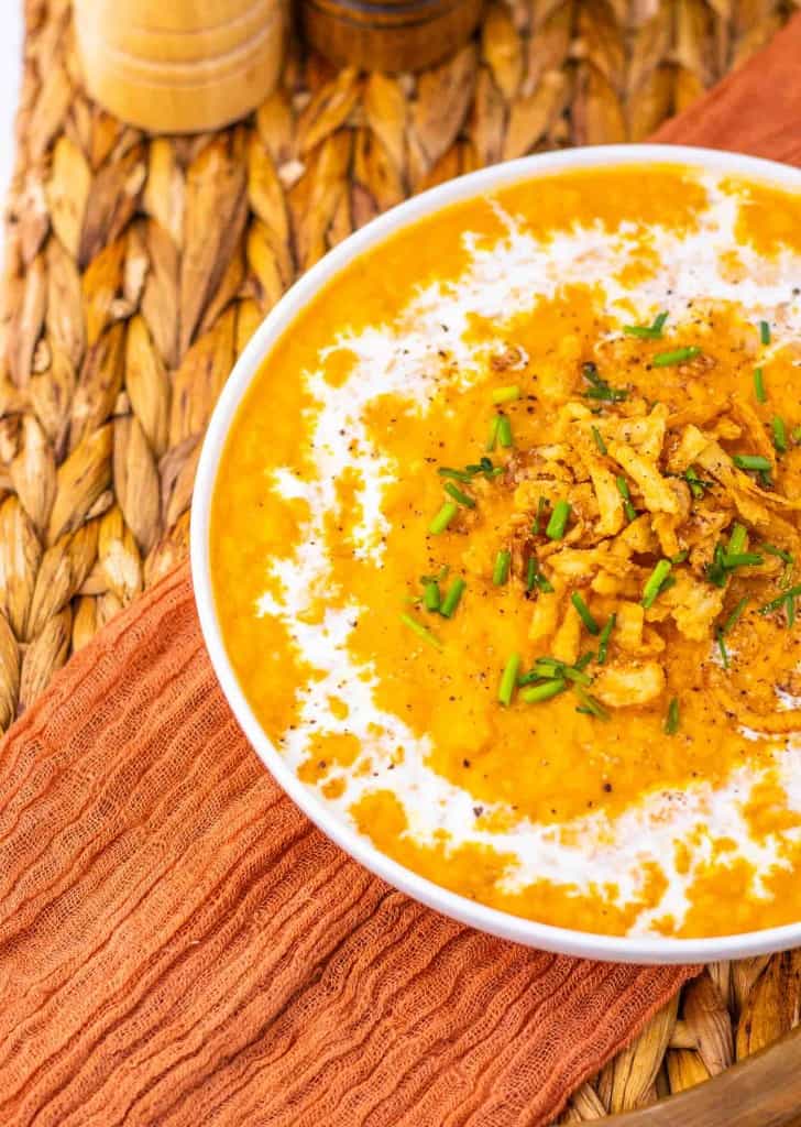 Bowl of creamy orange soup topped with crispy onions and chopped chives, placed on a woven mat with an orange cloth underneath.