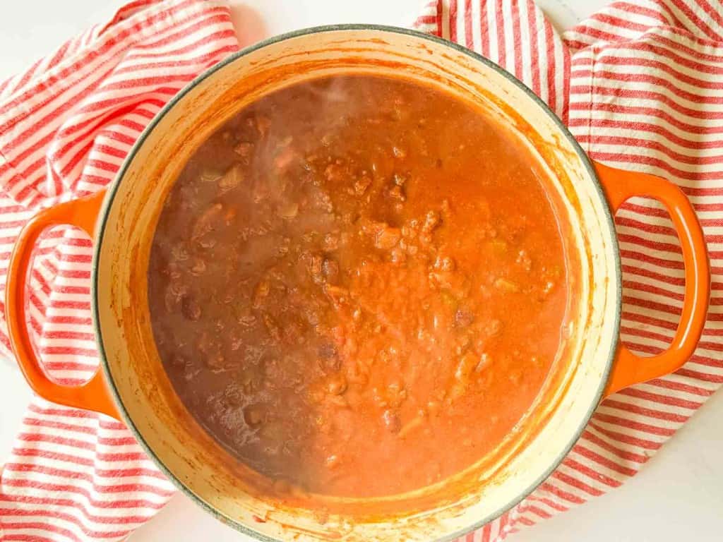 A pot of red chili with visible beans and meat simmers on a stove, surrounded by a red and white striped kitchen towel.