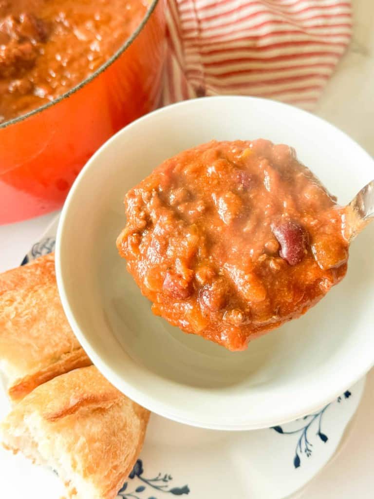 A spoonful of chili with beans held over a white bowl, with pieces of bread on the side and a red pot in the background.