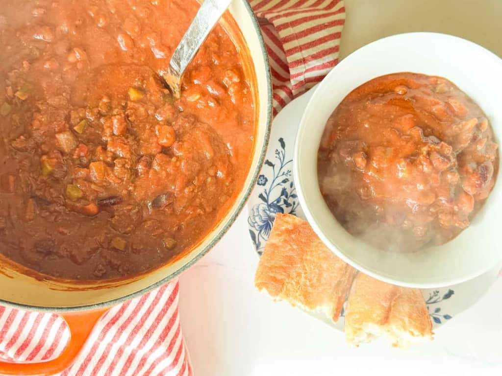 A pot and a bowl of chili with visible beans and meat are shown. The bowl is on a plate with two pieces of bread beside it. A red and white striped cloth is nearby.