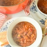 Bowl of chili with beans and vegetables on a floral plate, next to a piece of bread, spoon, and a red pot in the background.