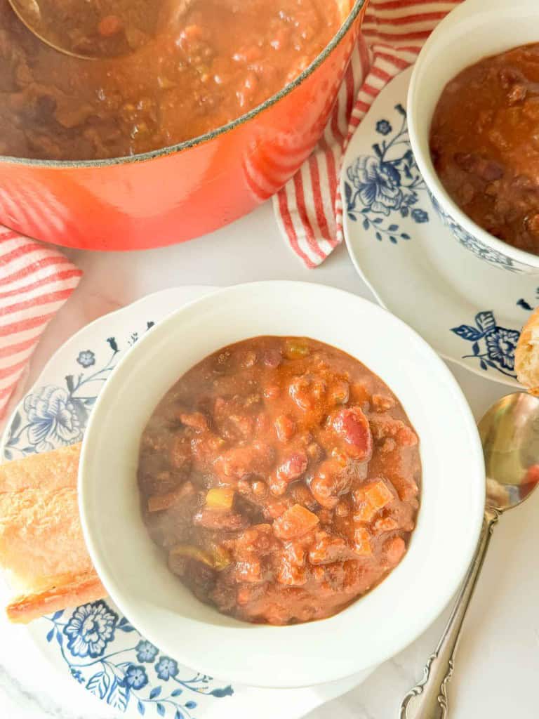 Bowl of chili with beans and vegetables on a floral plate, next to a piece of bread, spoon, and a red pot in the background.