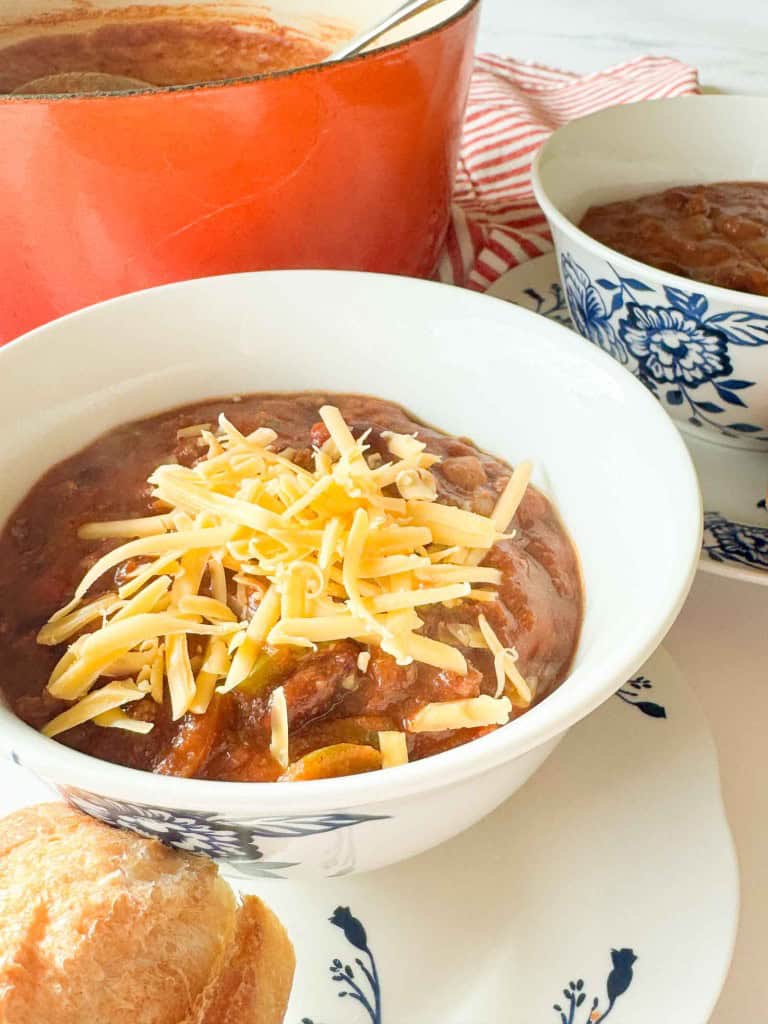 A white bowl of chili topped with shredded cheese sits on a floral-patterned plate with a bread roll, next to another bowl and a red pot in the background.