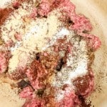 Raw ground beef in a pan topped with various spices and seasonings, ready to be mixed for cooking.