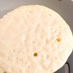 A buttermilk pancake is cooking in a pan with bubbles forming on the surface, indicating it is being cooked on one side.