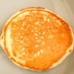 A single golden-brown buttermilk pancake with a slightly uneven edge sits on a light-colored surface.