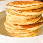 A stack of seven golden-brown buttermilk pancakes sits on a white plate, ready to be served.