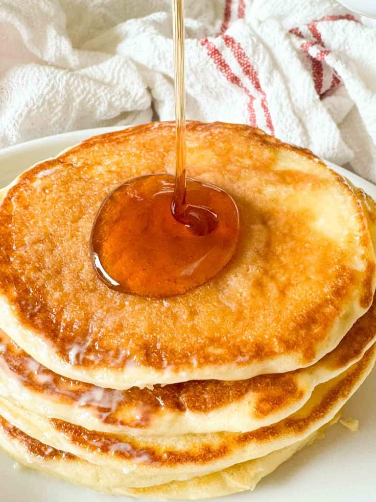 A stack of three golden buttermilk pancakes on a white plate with syrup being poured on top, in front of a white cloth.