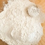 A pile of white flour on a wooden surface with a round object, perhaps destined to become fluffy buttermilk pancakes, partially covered in flour nearby.