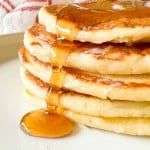 A stack of four golden buttermilk pancakes with syrup pouring down the side, served on a light-colored plate.