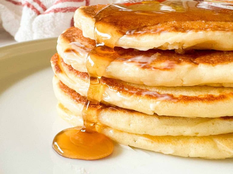 A stack of four golden buttermilk pancakes with syrup pouring down the side, served on a light-colored plate.