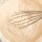 A metal whisk stirring thick, light beige buttermilk pancakes batter in a glass mixing bowl.