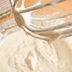 Close-up of a metal whisk covered in thick buttermilk pancake batter held above a glass bowl of more batter, with a measuring cup in the background.