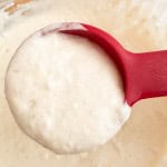 A red spoon holds thick buttermilk pancake batter above a mixing bowl filled with more batter.