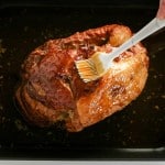 A hand uses a brush to add a rich, sweet coating to a Honey Glazed Ham in a black roasting pan.