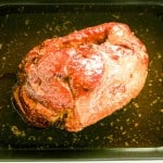 A honey glazed ham sits in a black roasting pan with juices at the bottom.
