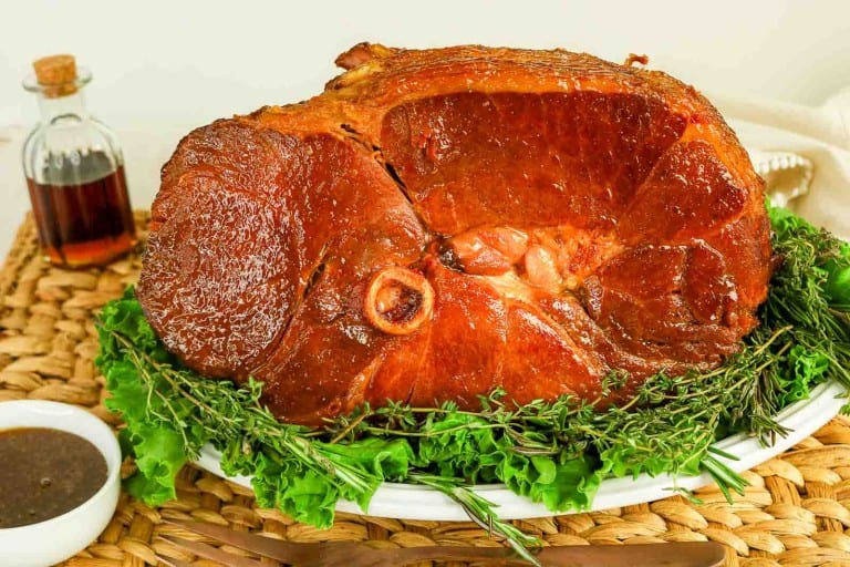 A Honey Glazed Ham sits on a white platter garnished with fresh herbs and greens, with a small bottle of sauce and a bowl of dipping sauce nearby.