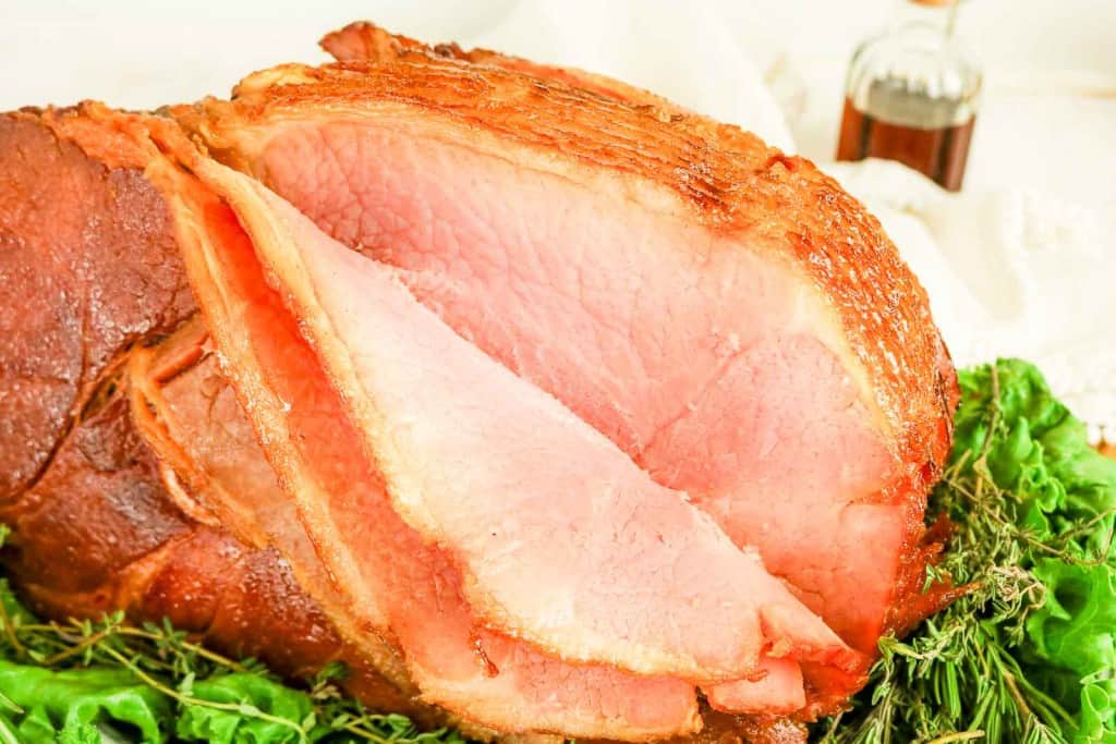 Close-up of sliced Honey Glazed Ham on a bed of greens with a small bottle of liquid in the background.