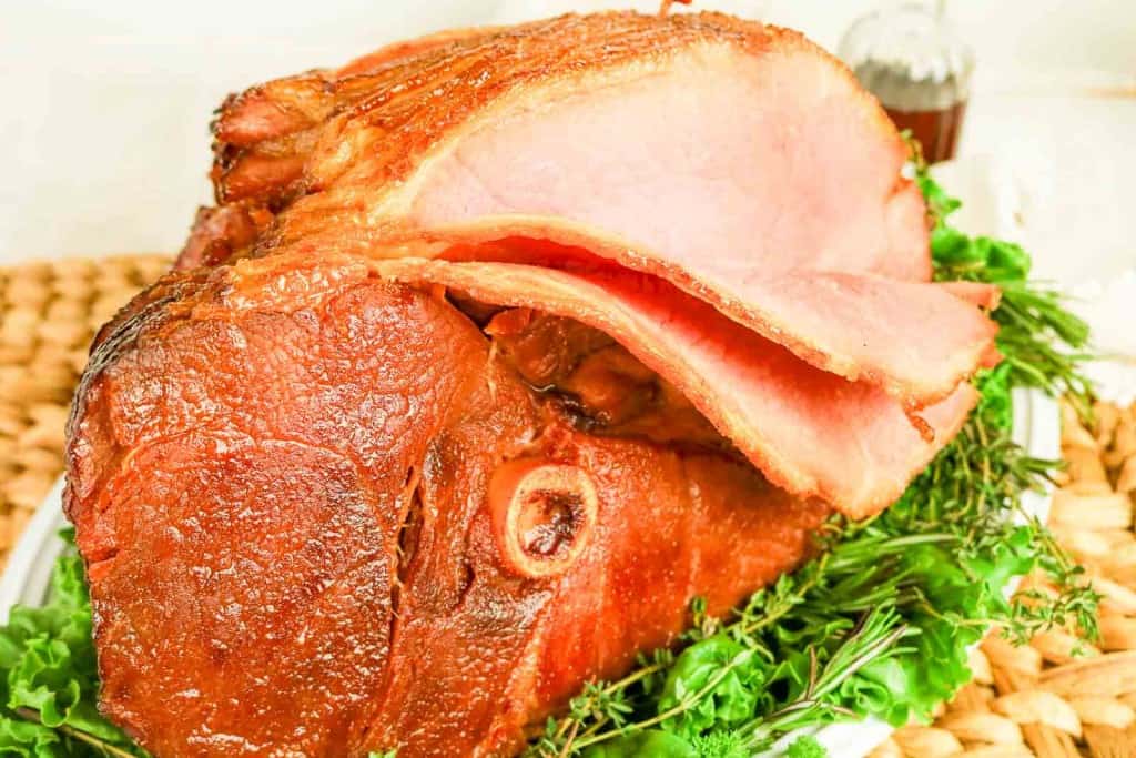 A Honey Glazed Ham with several slices cut, served on a bed of leafy greens and herbs.