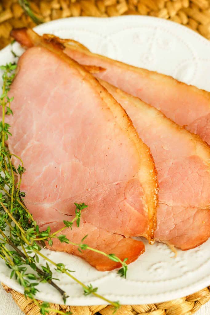 Two slices of honey glazed ham on a white plate, garnished with sprigs of fresh thyme.