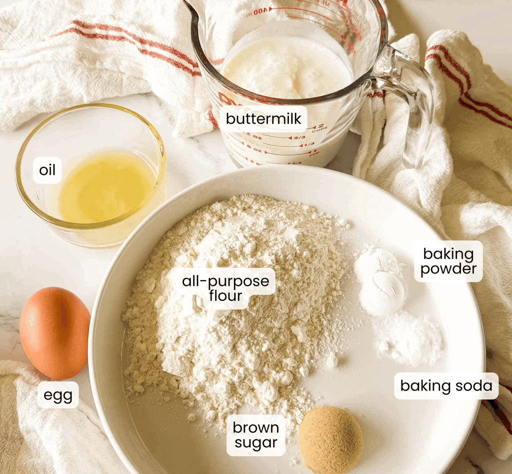 A bowl with flour, baking powder, baking soda, and brown sugar; a cup of buttermilk for fluffy buttermilk pancakes; a small bowl of oil; and an egg on a light surface, each ingredient labeled.