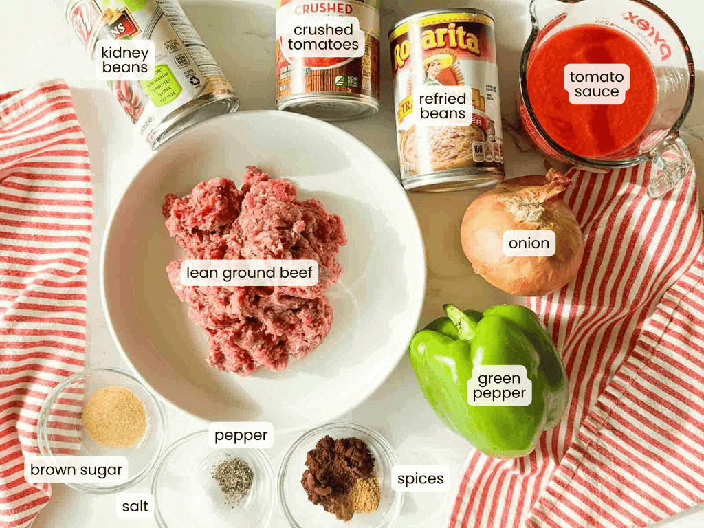 Ingredients for beef chili are arranged on a white surface, including ground beef, green pepper, onion, various beans, tomato sauce, crushed tomatoes, and seasonings in small bowls.