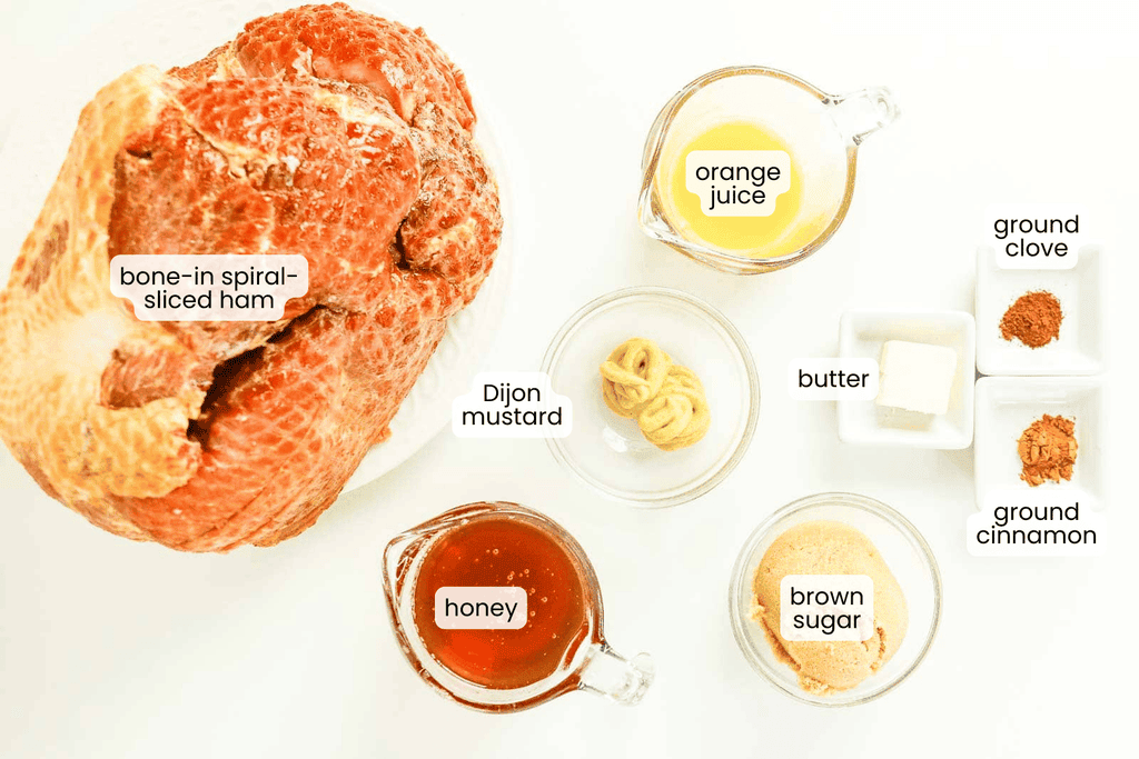 Bone-in spiral-sliced Honey Glazed Ham, honey, Dijon mustard, orange juice, butter, brown sugar, ground cinnamon, and ground clove are arranged on a white surface in separate bowls.