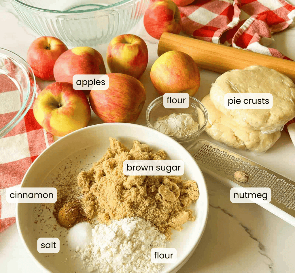 Baking ingredients for an old fashioned apple pie are labeled on a counter: apples, flour, brown sugar, cinnamon, salt, nutmeg, and pie crusts, with a rolling pin, mixing bowls, and a red checkered towel.