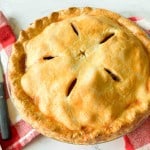 Whole baked old fashioned apple pie with golden brown crust and five slits on top, placed on a red and white checkered cloth next to a pie server and knife.