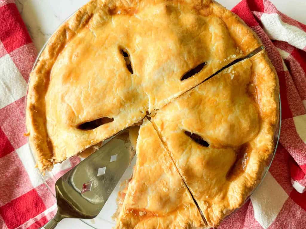 A baked old fashioned apple pie with a golden crust sits on a red and white checkered cloth, with three slices cut and a pie server beside it.