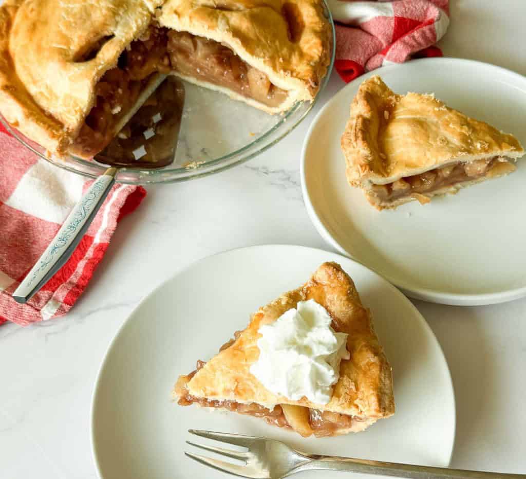 A sliced old fashioned apple pie in a glass dish with two pieces on white plates, one topped with whipped cream. A fork and a pie server are next to the plates.