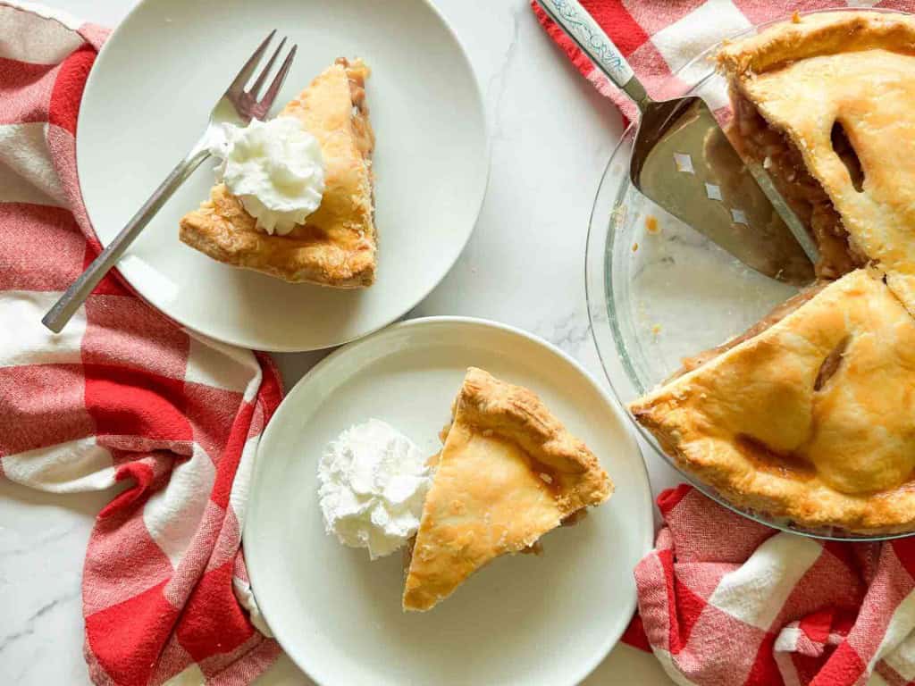Two plates with slices of old fashioned apple pie and whipped cream sit on a table next to a pie dish with more pie, with a red and white checkered cloth nearby.