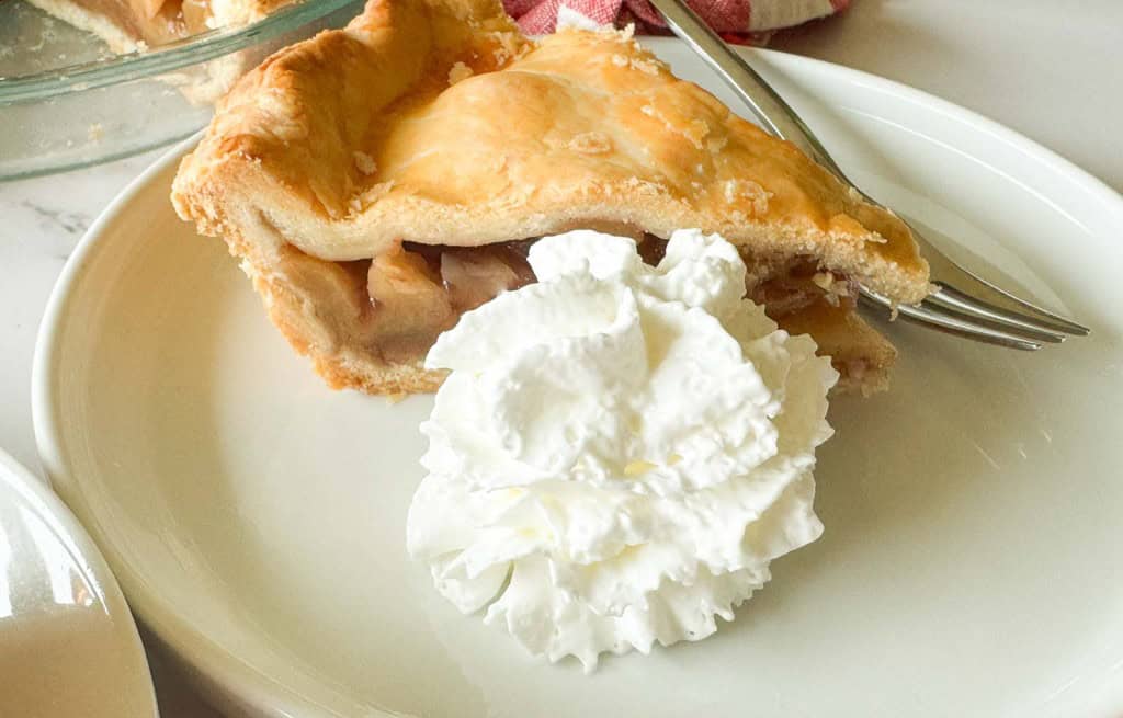A slice of old fashioned apple pie with a flaky crust sits on a white plate next to a swirl of whipped cream and a fork.