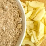 A bowl of crumbly topping sits next to a pile of peeled, sliced apples on a wooden surface, ready to become the heart of an old fashioned apple pie.