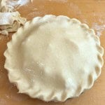 Unbaked old fashioned apple pie with crimped edges on a floured wooden surface, with scraps of dough to the side.