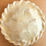 Unbaked old fashioned apple pie with a crimped edge and five slits in the top crust sits on a wooden surface.