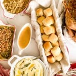 Thanksgiving meal with bread rolls, mashed potatoes with herbs, gravy, green bean casserole, stuffing, and roasted turkey on a white table.
