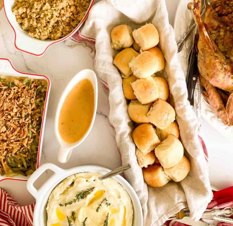 Thanksgiving meal with bread rolls, mashed potatoes with herbs, gravy, green bean casserole, stuffing, and roasted turkey on a white table.