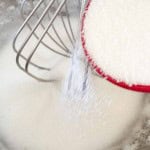 A red measuring cup pours granulated sugar into a mixing bowl next to a wire whisk.