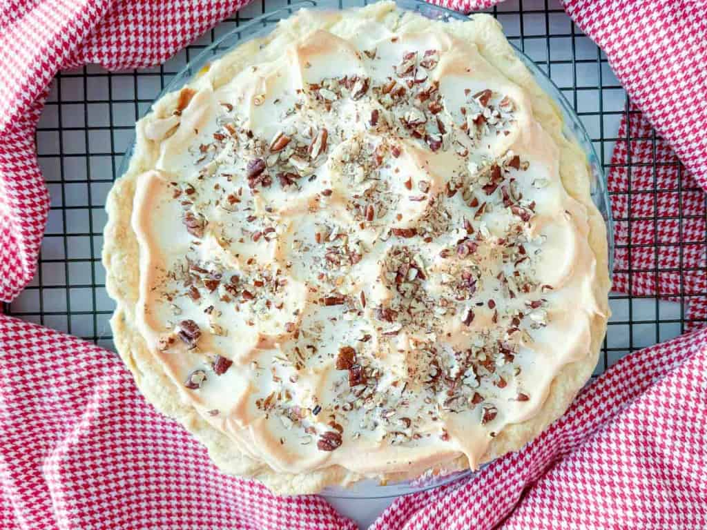 Meringue pie topped with chopped pecans on a wire cooling rack over a red and white checkered cloth.