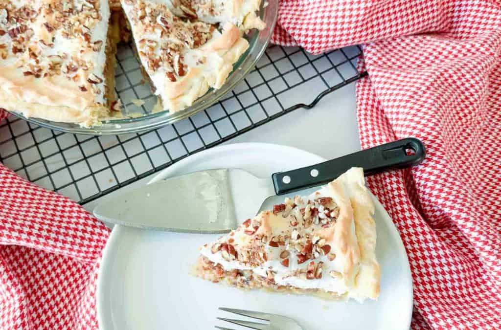 A slice of pecan meringue pie on a white plate with a pie server, next to the remaining pie in a glass dish on a cooling rack.