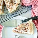 A slice of pecan meringue pie on a white plate with a pie server, next to the remaining pie in a glass dish on a cooling rack.