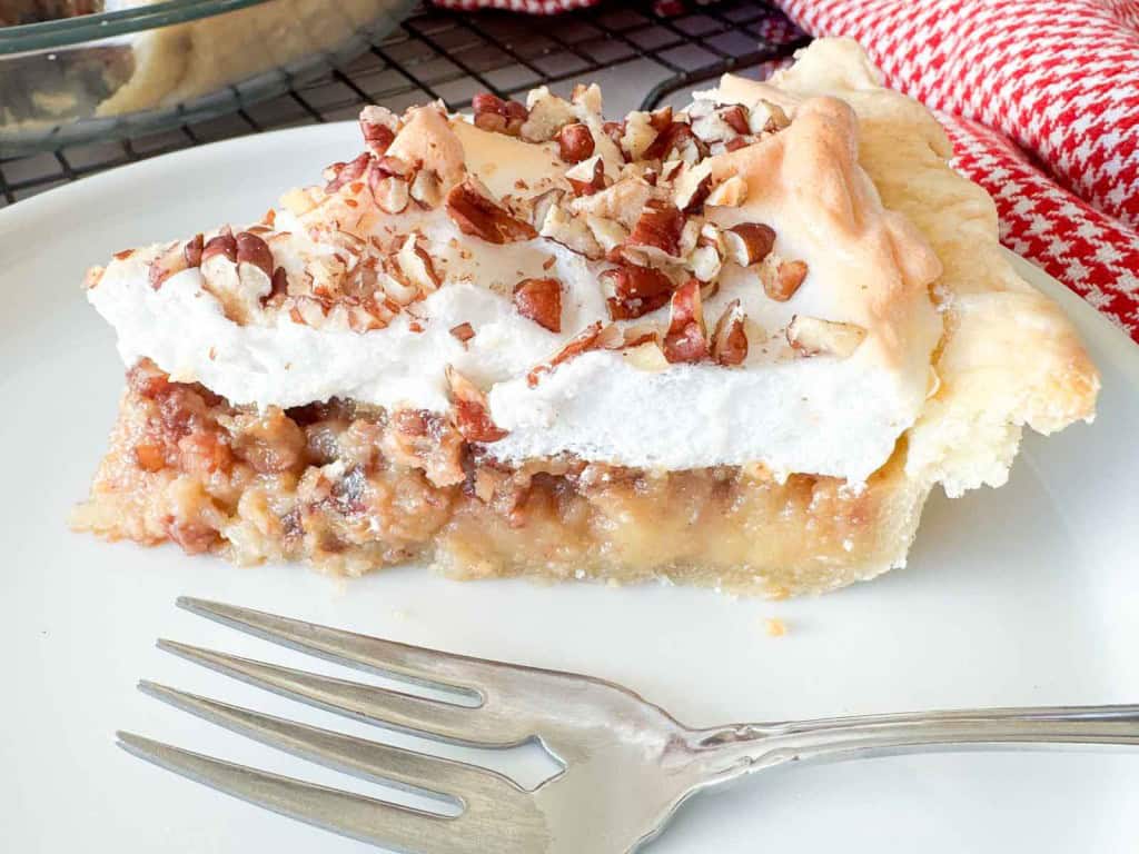A slice of pie with a dense filling, topped with meringue and chopped pecans, on a white plate with a fork.