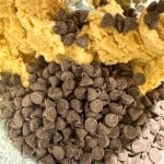 A pile of dark chocolate chips sitting atop unmixed cookie dough in a mixing bowl.