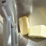 Two sticks of butter in a stainless steel stand mixer bowl next to the paddle attachment.