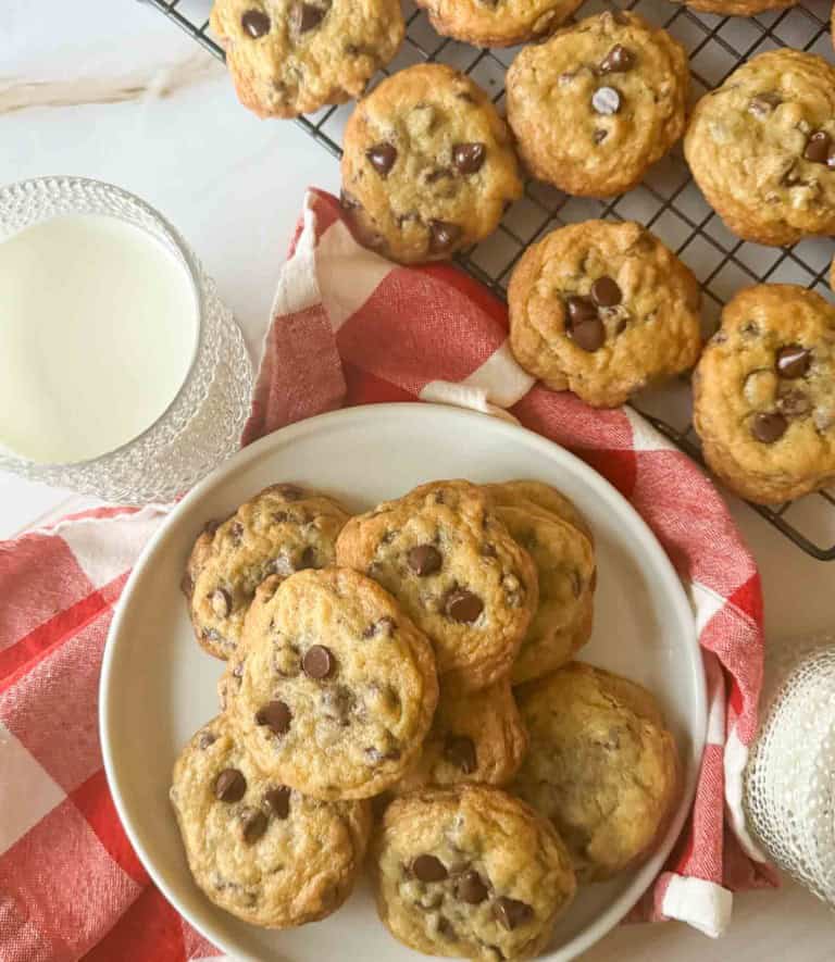 A plate piled with chocolate chip cookies next to a wire cooling rack of more cookies and a glass of milk.